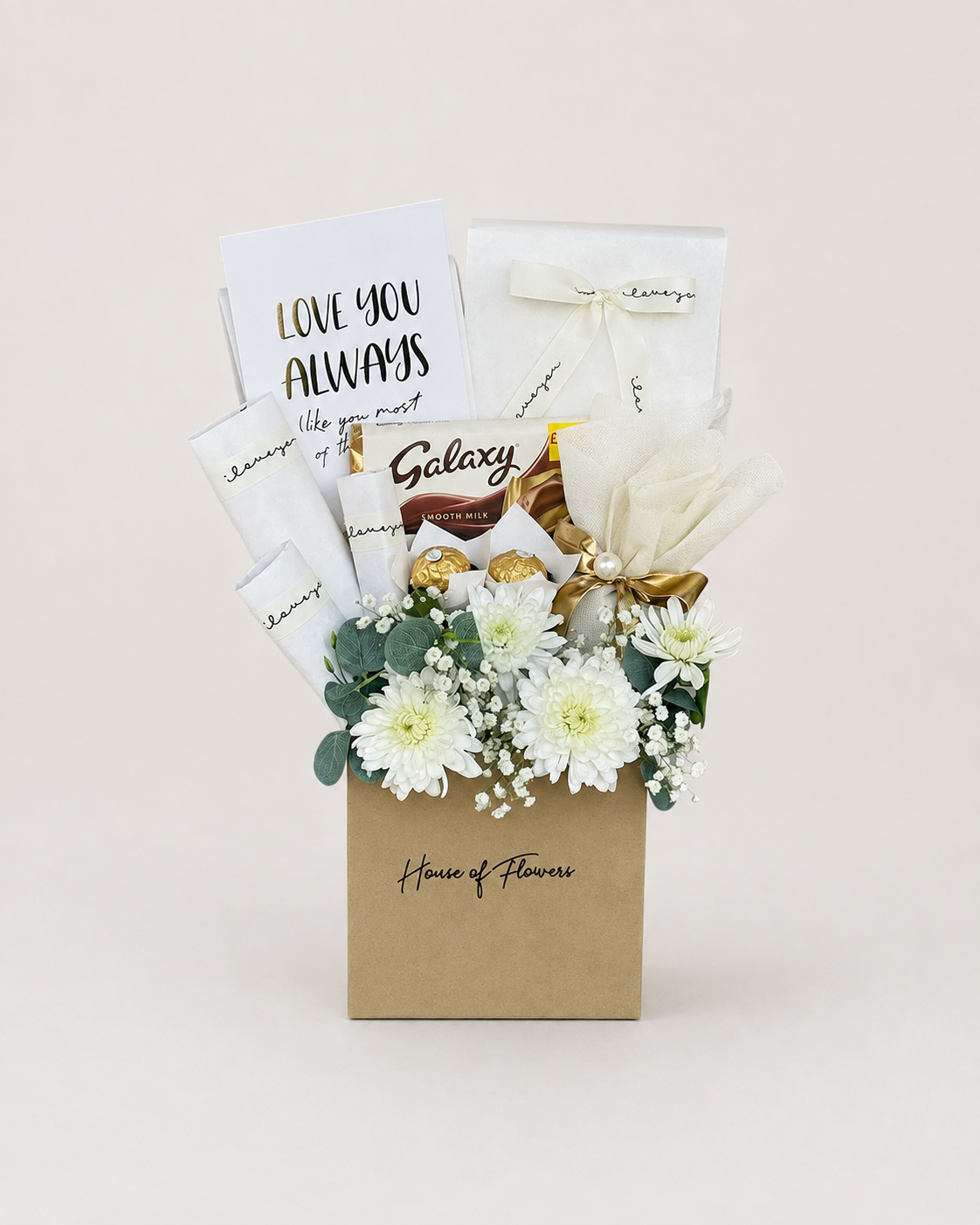 A gourmet gift basket from House of Flowers featuring white flowers, chocolates, and gift cards. The kraft paper basket is printed with &