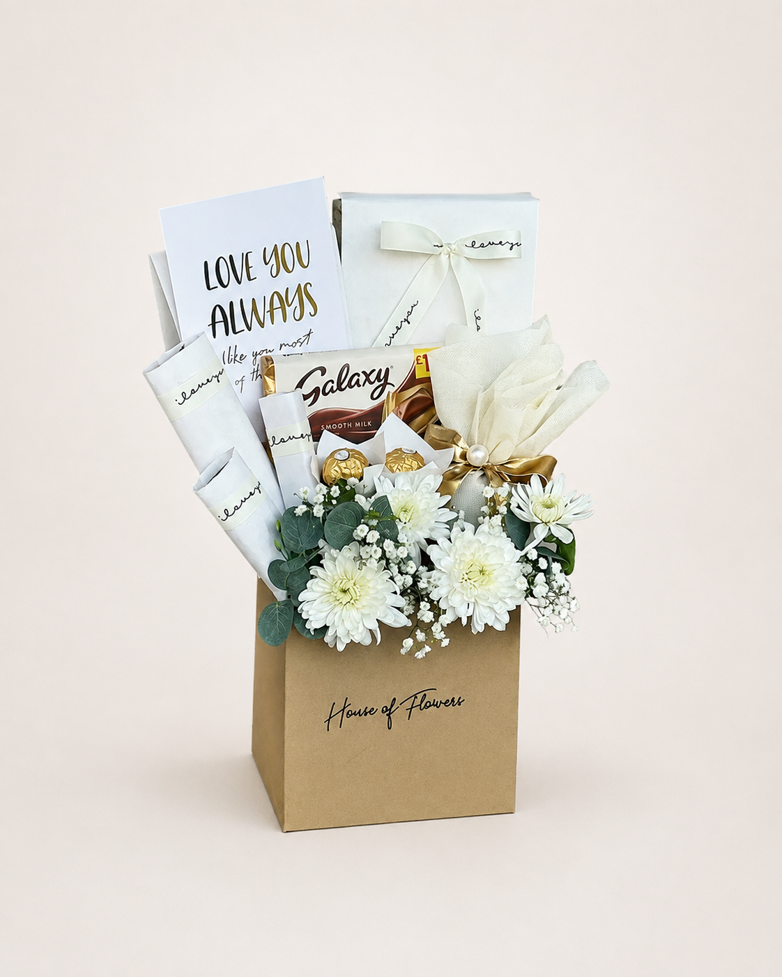 A gourmet gift box from House of Flowers, featuring a brown kraft paper bag filled with white flowers, chocolates, and gift cards, set against a solid light beige background.