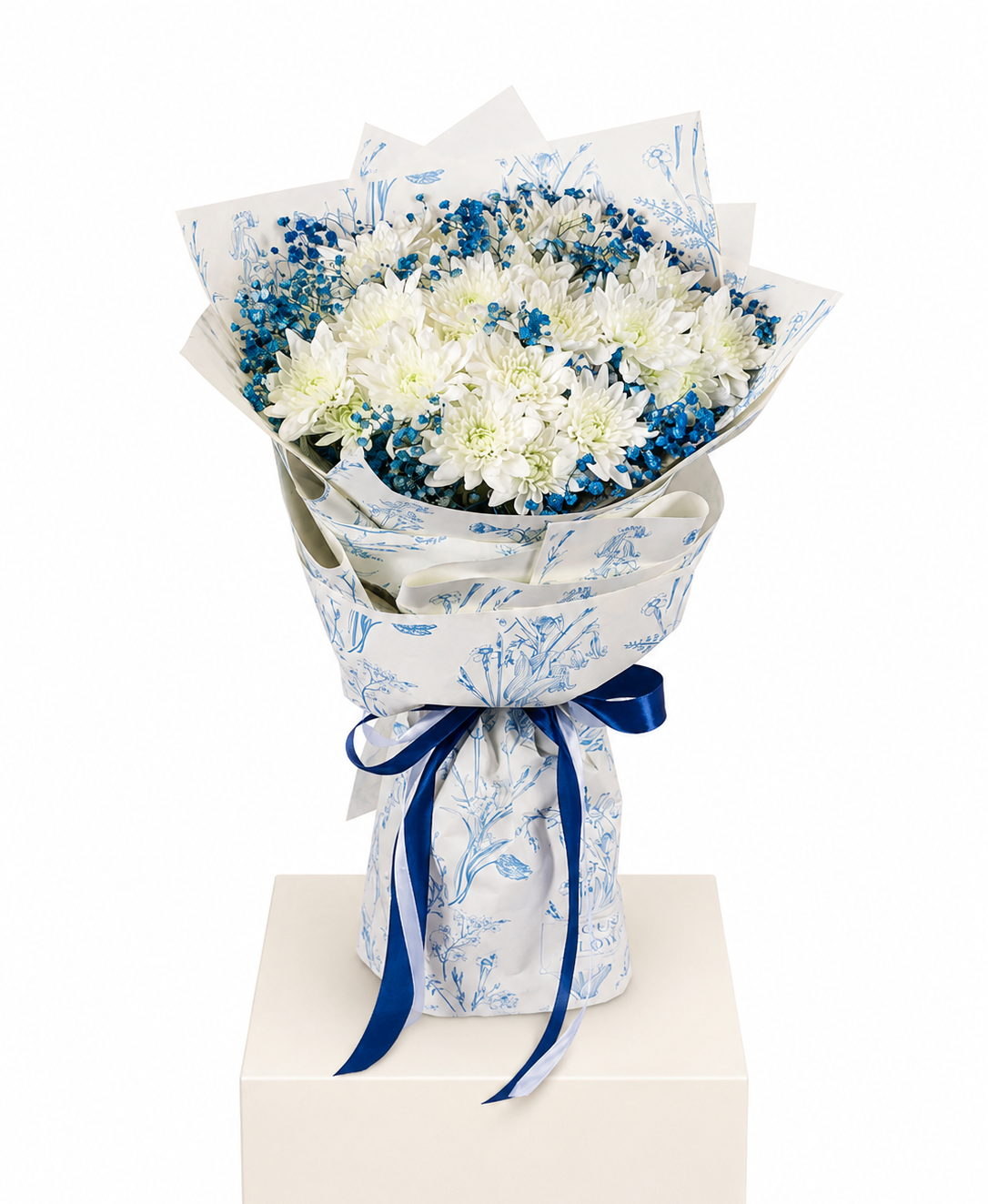 A bouquet of white and blue flowers wrapped in white paper with a blue botanical pattern and tied with a dark blue ribbon, displayed on a white pedestal against a white background.