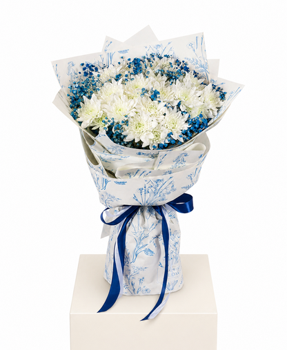A bouquet of white and blue flowers wrapped in white paper with a blue botanical pattern and tied with a dark blue ribbon, displayed on a white pedestal against a white background.