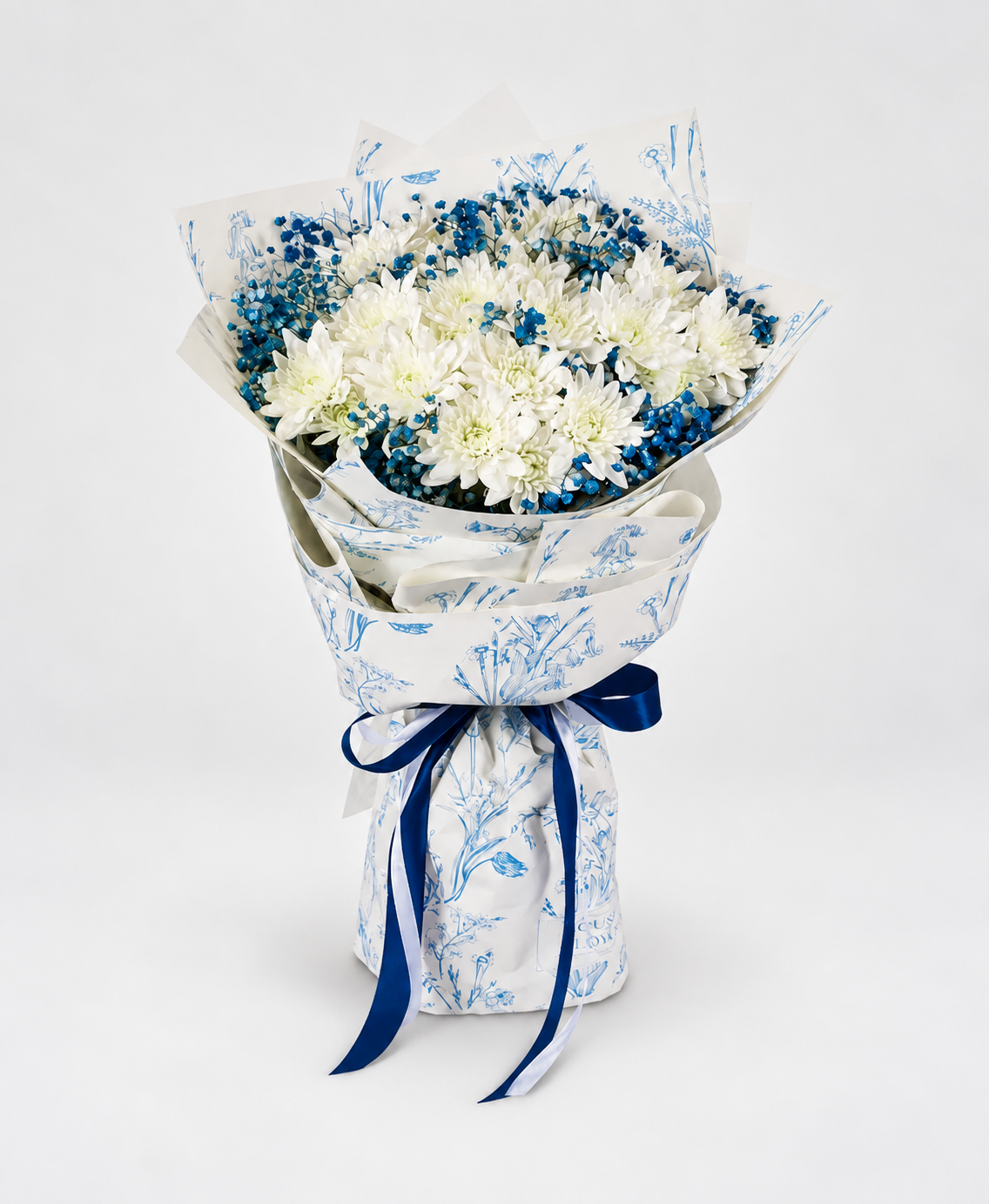 A bouquet of white and blue flowers wrapped in white paper with a blue botanical pattern and tied with a dark blue ribbon, displayed on a white pedestal against a white background.