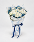 A bouquet of white and blue flowers wrapped in white paper with a blue botanical pattern and tied with a dark blue ribbon, displayed on a white pedestal against a white background.