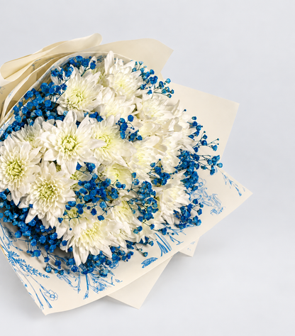 A bouquet of white and blue flowers wrapped in white paper with a blue botanical pattern and tied with a dark blue ribbon, displayed on a white pedestal against a white background.