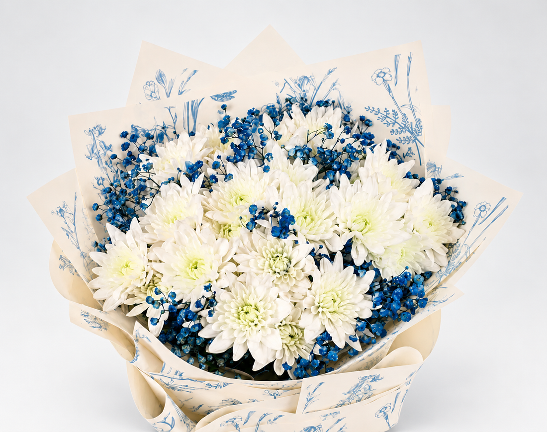 A bouquet of white and blue flowers wrapped in white paper with a blue botanical pattern and tied with a dark blue ribbon, displayed on a white pedestal against a white background.