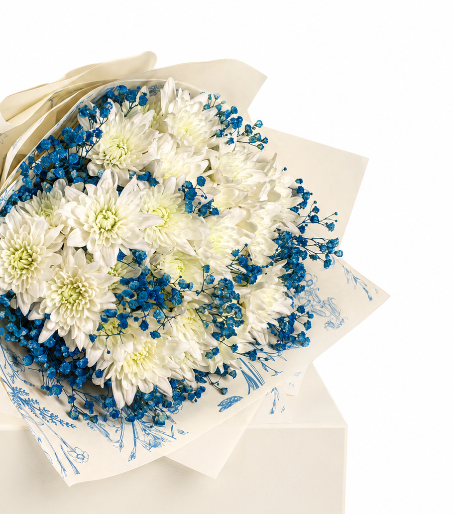 A bouquet of white chrysanthemums with blue babys breath flowers, wrapped in cream-colored paper with blue floral patterns, displayed on a cream gift box against a white background.