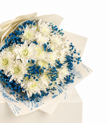 A bouquet of white chrysanthemums with blue babys breath flowers, wrapped in cream-colored paper with blue floral patterns, displayed on a cream gift box against a white background.