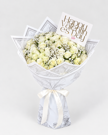 A bouquet of white and pale yellow roses wrapped in white patterned paper with a cream ribbon, topped with a card that reads &