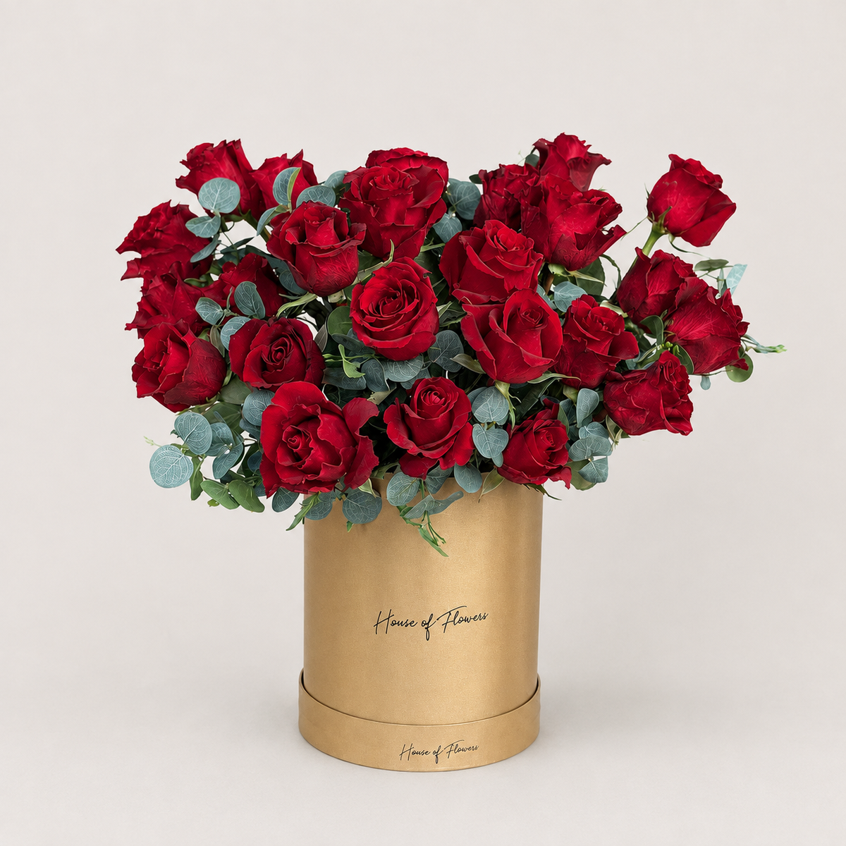 A bouquet of red roses with green eucalyptus stems arranged in a white cylindrical box printed with the gold script &