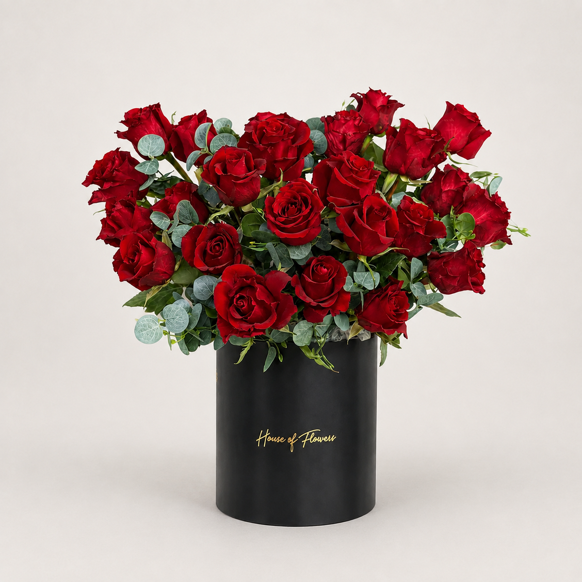 A bouquet of red roses with green eucalyptus stems arranged in a white cylindrical box printed with the gold script &