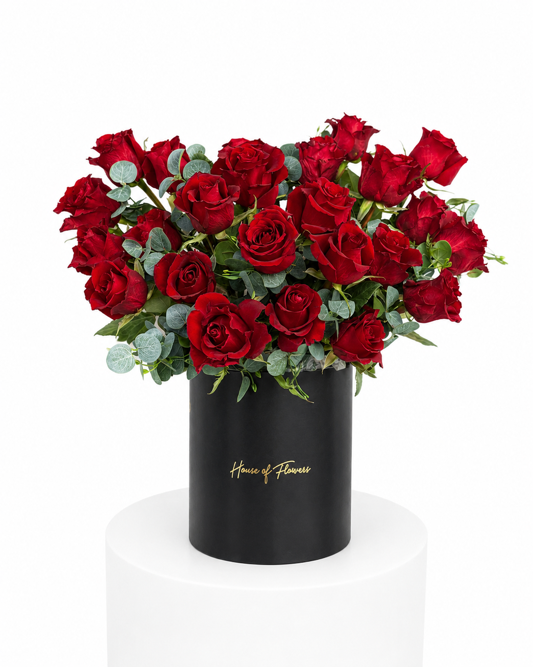 A vibrant bouquet of red roses with green foliage arranged in a black cylindrical box with gold &