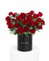 A vibrant bouquet of red roses with green foliage arranged in a black cylindrical box with gold &