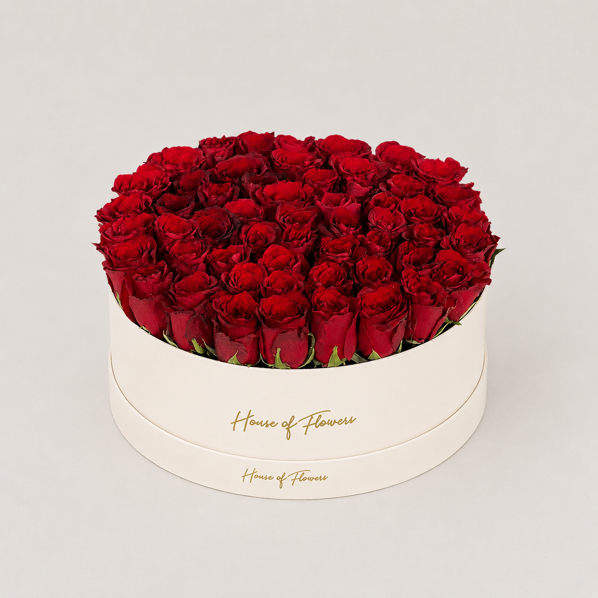A round white gift box filled with a dense arrangement of deep red roses, photographed on a clean light gray background. The box is printed with the brand name &