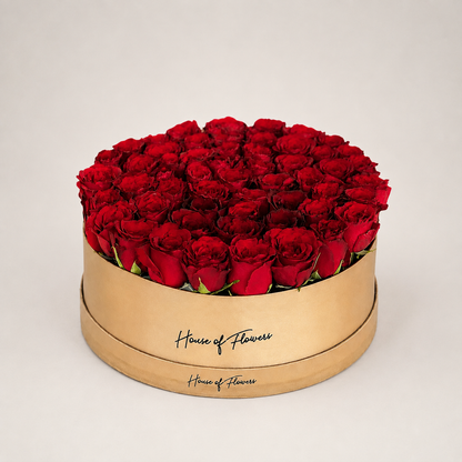 A round gift box filled with red roses, featuring a gold-colored wooden base printed with the brand name &