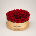 A round gift box filled with red roses, featuring a gold-colored wooden base printed with the brand name &