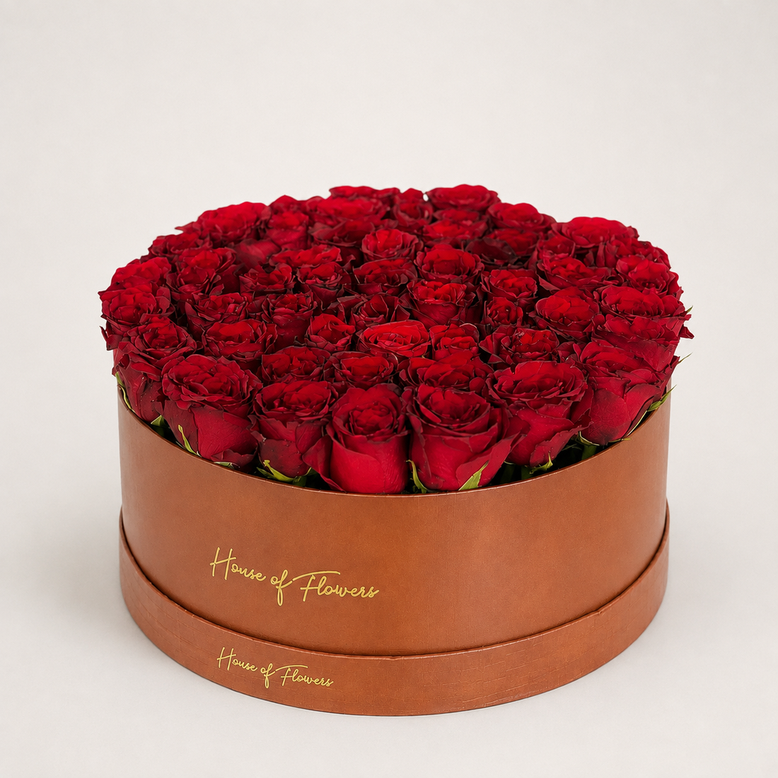 A round gift box filled with deep red roses, displayed on a white pedestal against a white background. The box is cream-colored with gold &
