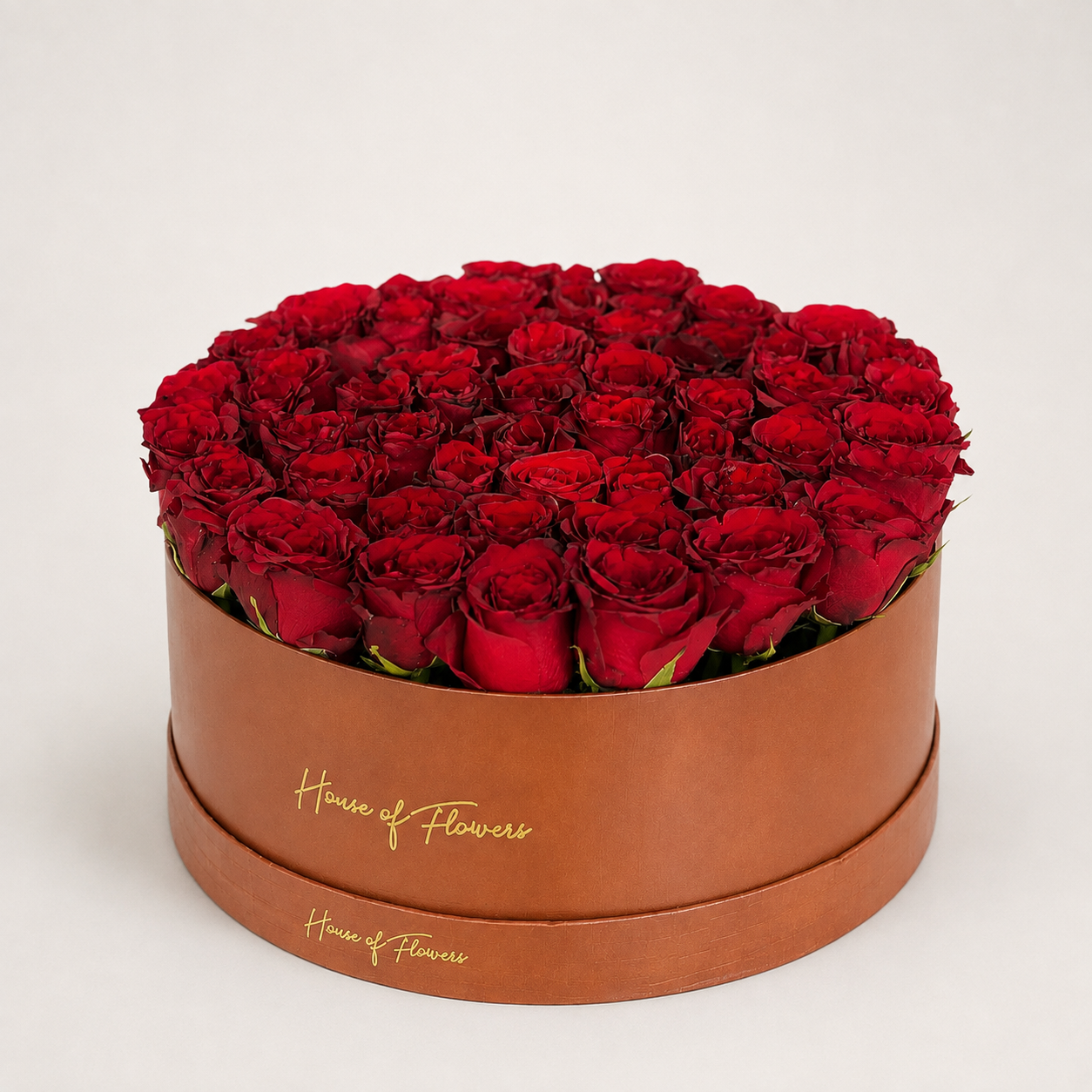 A round gift box filled with deep red roses, displayed on a white pedestal against a white background. The box is cream-colored with gold &