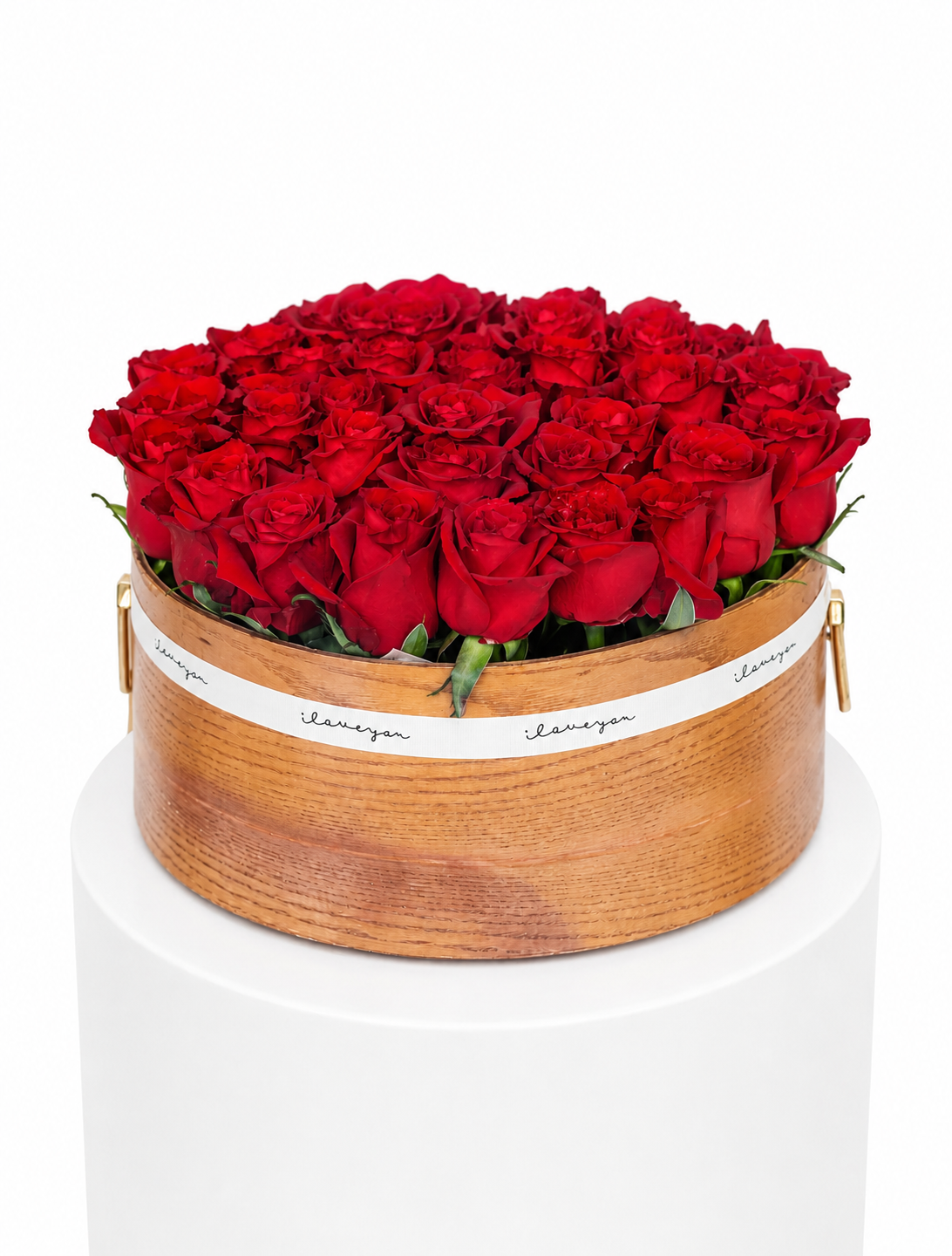 A round wooden box filled with red roses, wrapped with a white ribbon printed with the brand name &