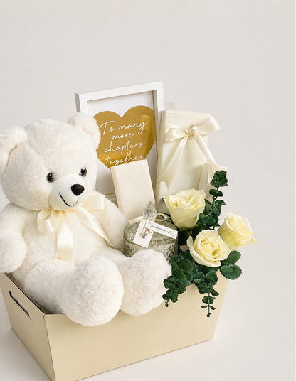 A white plush teddy bear wearing a cream ribbon sits in a cream gift box alongside pale yellow roses, a wrapped gift with a bow, a small card, and a framed heart sign that reads &