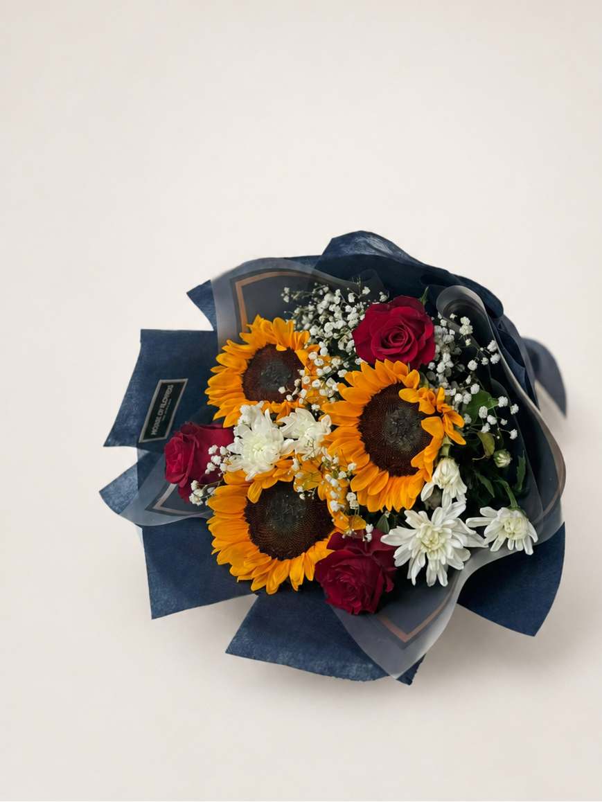 Bouquet of sunflowers, roses, and other flowers wrapped in dark paper with a card on a white surface.