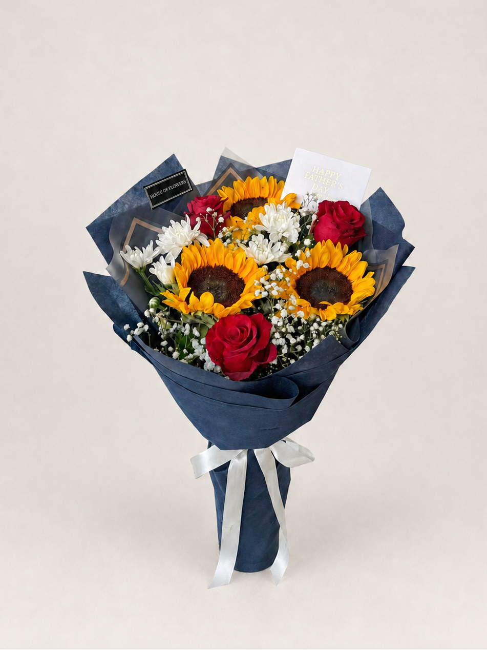 Bouquet of sunflowers, roses, and other flowers wrapped in dark paper with a card on a white surface.