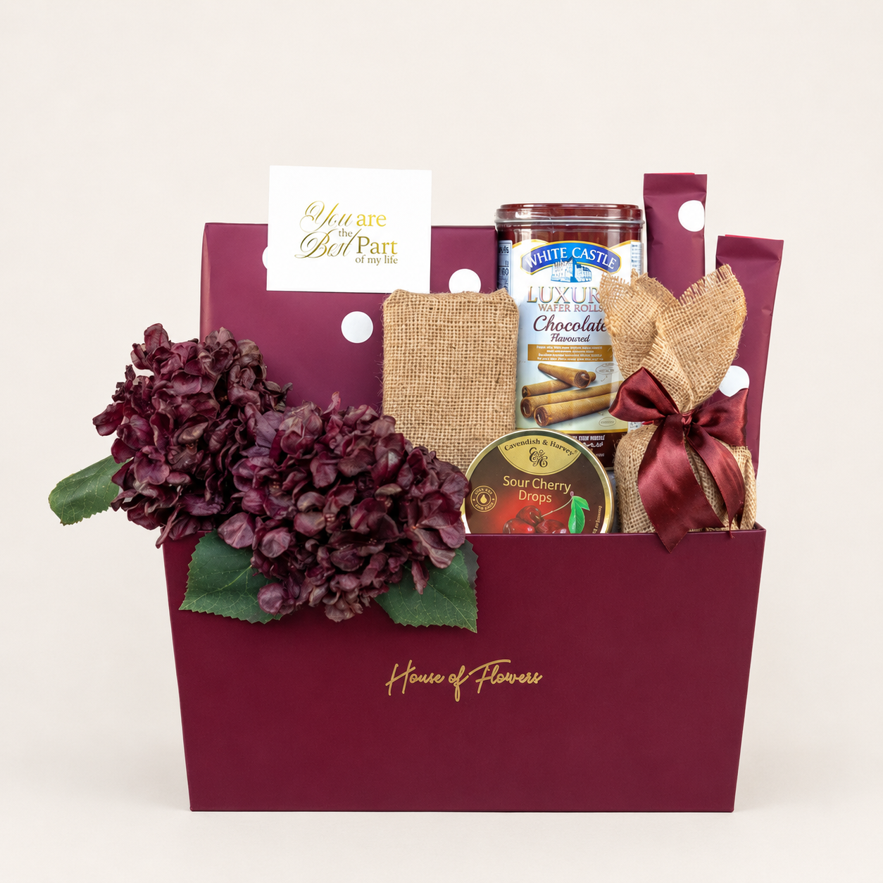 A gourmet gift basket from House of Flowers, featuring a deep burgundy paper bag filled with a large dark purple hydrangea, a burlap-wrapped item tied with a burgundy ribbon, a canister of White Castle Luxury Chocolate, and a small jar of Cherry Limeade, all set against a plain off-white background.