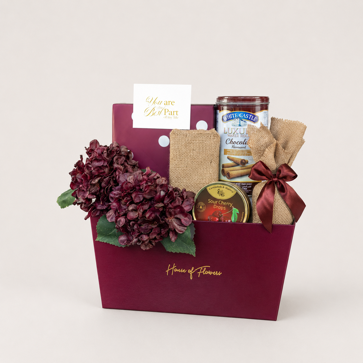 A gourmet gift basket from House of Flowers by White Castle, featuring a deep burgundy box with gold lettering, filled with a bouquet of dark burgundy hydrangeas, a burlap-wrapped White Castle chocolate bar, a jar of White Castle Muffins, and a small tin of White Castle Sea Cherry Cinnamon Scones, complete with a &