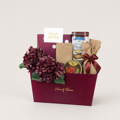 A gourmet gift basket from House of Flowers by White Castle, featuring a deep burgundy box with gold lettering, filled with a bouquet of dark burgundy hydrangeas, a burlap-wrapped White Castle chocolate bar, a jar of White Castle Muffins, and a small tin of White Castle Sea Cherry Cinnamon Scones, complete with a &
