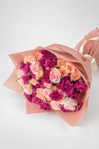 A bouquet of flowers consisting of roses and chrysanthemums in shades of pink and purple, wrapped in pink paper with a ribbon and a card that reads &