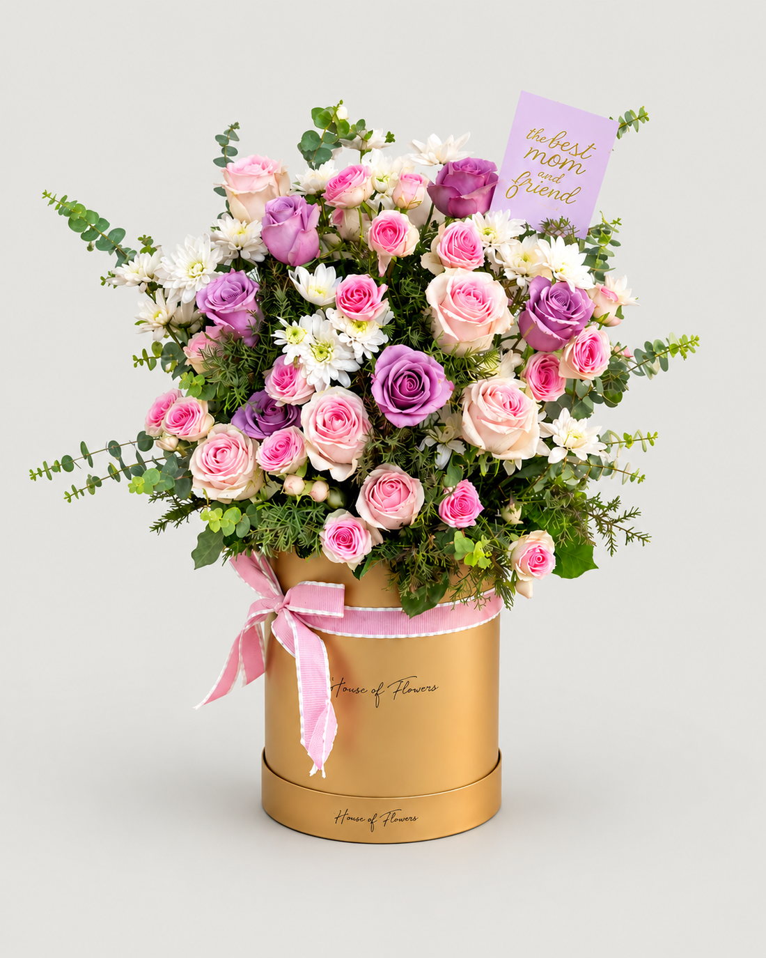 A floral gift arrangement featuring pink and purple roses, white chrysanthemums, and green foliage in a round gold box with a pink ribbon, set against a light gray background. A small lavender card reads &