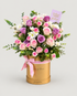 A floral gift arrangement featuring pink and purple roses, white chrysanthemums, and green foliage in a round gold box with a pink ribbon, set against a light gray background. A small lavender card reads &