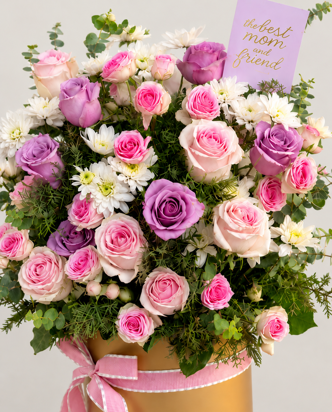 A vibrant floral arrangement featuring pink and purple roses, white chrysanthemums, and green foliage in a gold-colored box tied with a pink ribbon, accompanied by a lavender card that reads &