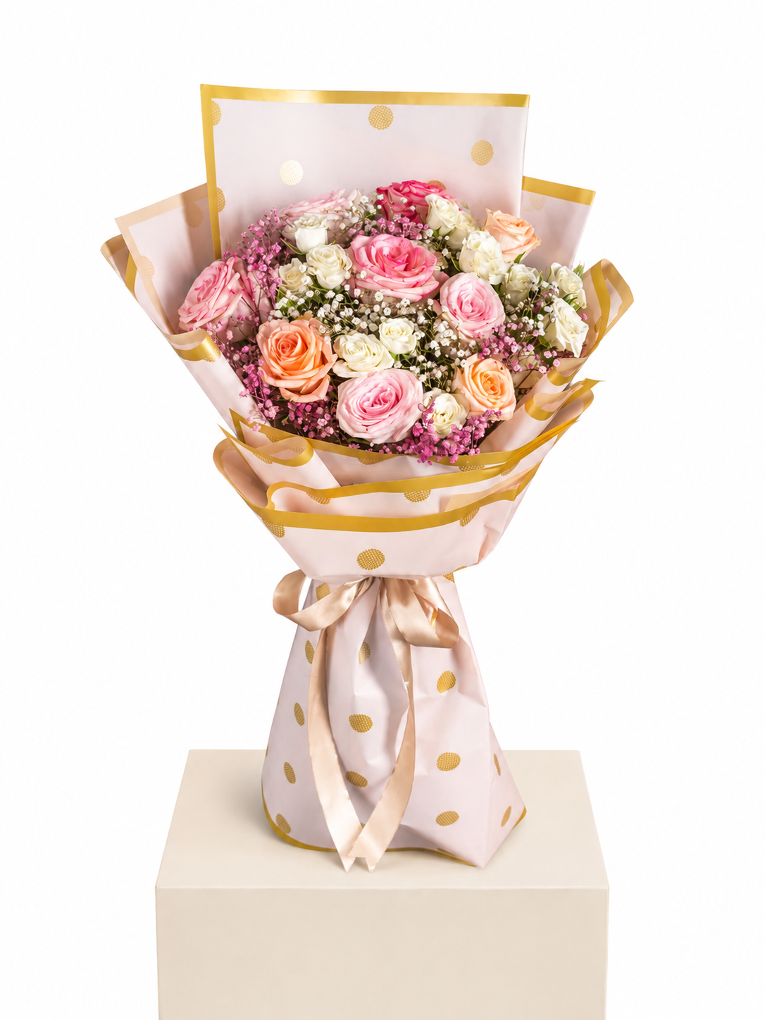 A bouquet of pink, peach, and white roses wrapped in light pink polka-dot paper with gold trim, tied with a satin ribbon, displayed on a cream pedestal against a white background.