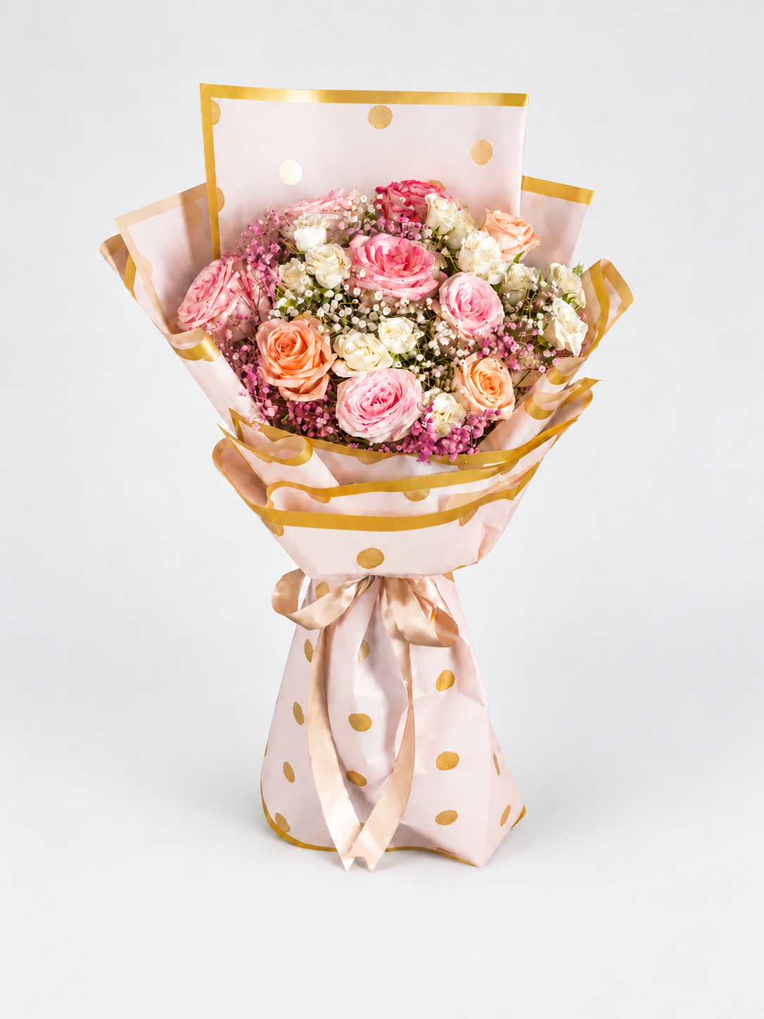 A bouquet of pink, peach, and white roses wrapped in light pink polka-dot paper with gold trim, tied with a satin ribbon, displayed on a cream pedestal against a white background.