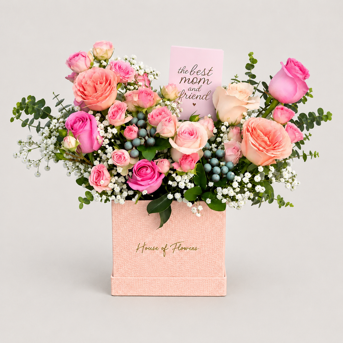 A floral gift arrangement in a pink box from House of Flowers, featuring pink and peach roses, green foliage, small white flowers, blue berries, and a card that reads &