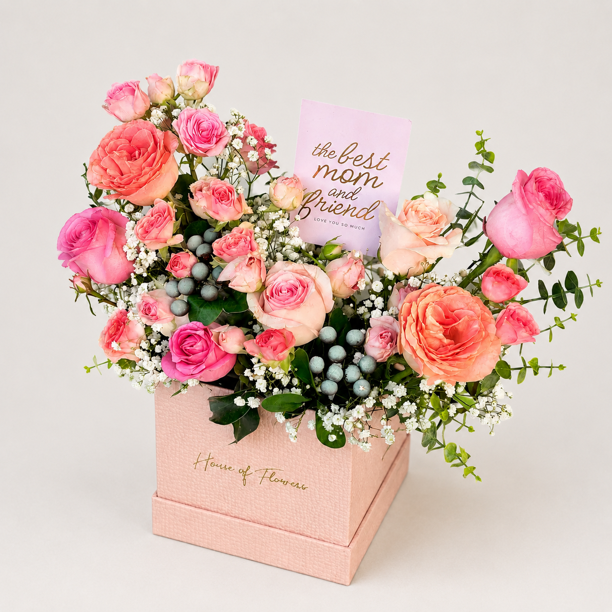 A floral gift arrangement in a pink box from House of Flowers, featuring pink and peach roses, green foliage, small white flowers, blue berries, and a card that reads &