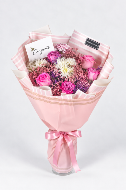 A bouquet of flowers including roses and chrysanthemums in purple, pink, and white colors, with a pink veil wrapped around the stems, tied with a pink ribbon.