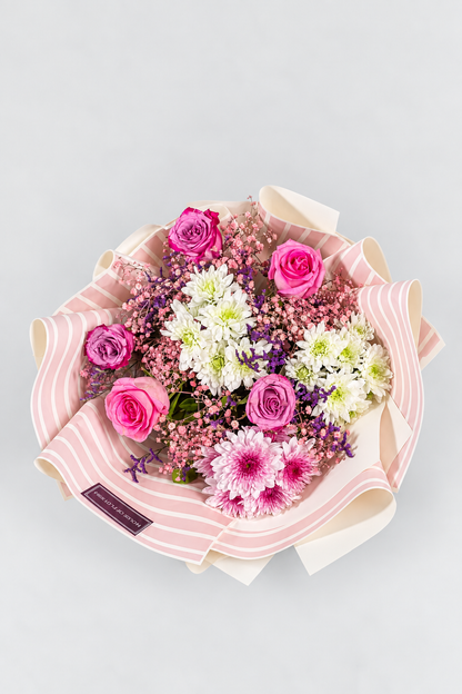 A bouquet of flowers including roses and chrysanthemums in purple, pink, and white colors, with a pink veil wrapped around the stems, tied with a pink ribbon.