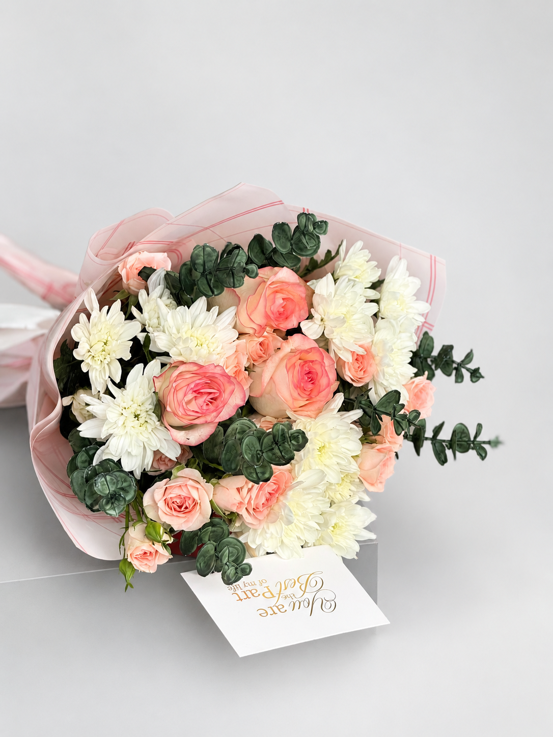 A floral bouquet with pink and white flowers, green foliage, and a small card that reads &