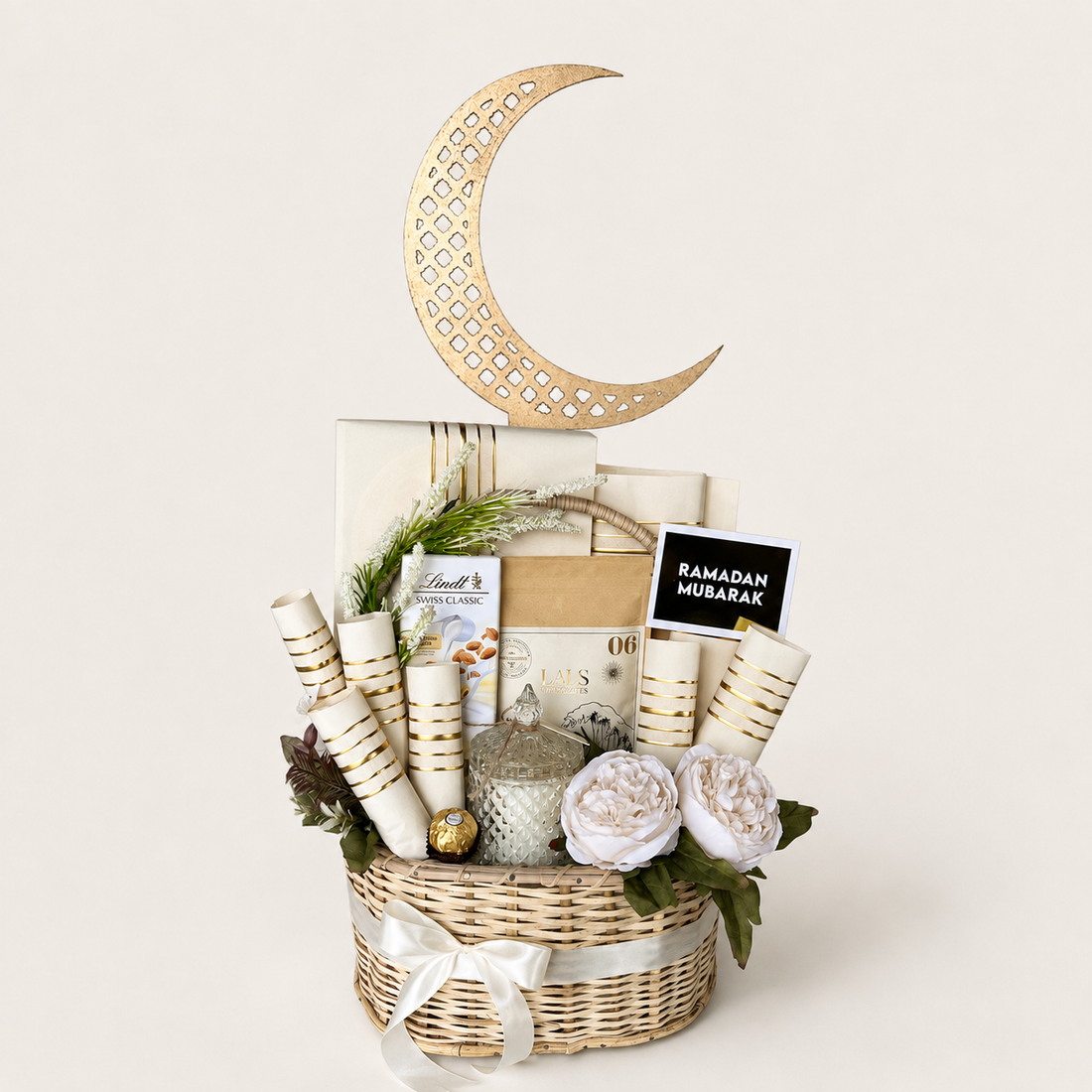 A Ramadan gift basket arrangement in a woven wicker basket, featuring a large gold crescent moon decoration, rolled white and gold mubarak cards, wrapped gift boxes, a small silver lantern, faux white flowers, and a black sign reading &