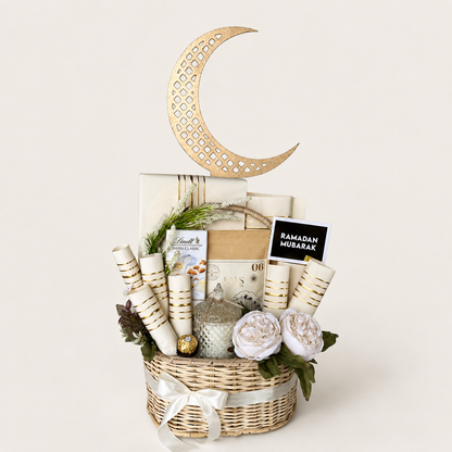 A Ramadan gift basket arrangement in a woven wicker basket, featuring a large gold crescent moon decoration, rolled white and gold mubarak cards, wrapped gift boxes, a small silver lantern, faux white flowers, and a black sign reading &