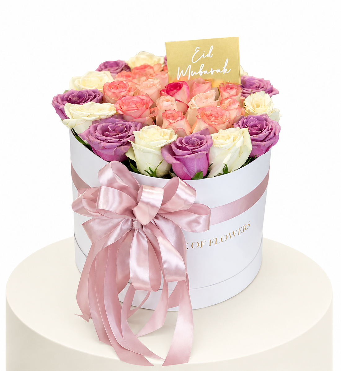 A round white gift box filled with assorted roses in pastel shades of pink, purple, peach, and cream, tied with a large pink satin bow. A small gold card reading &