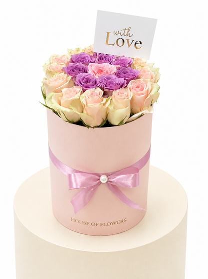 A round pink flower box filled with pale pink and purple roses, tied with a pink ribbon and topped with a small card that reads &
