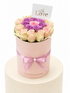 A round pink flower box filled with pale pink and purple roses, tied with a pink ribbon and topped with a small card that reads &