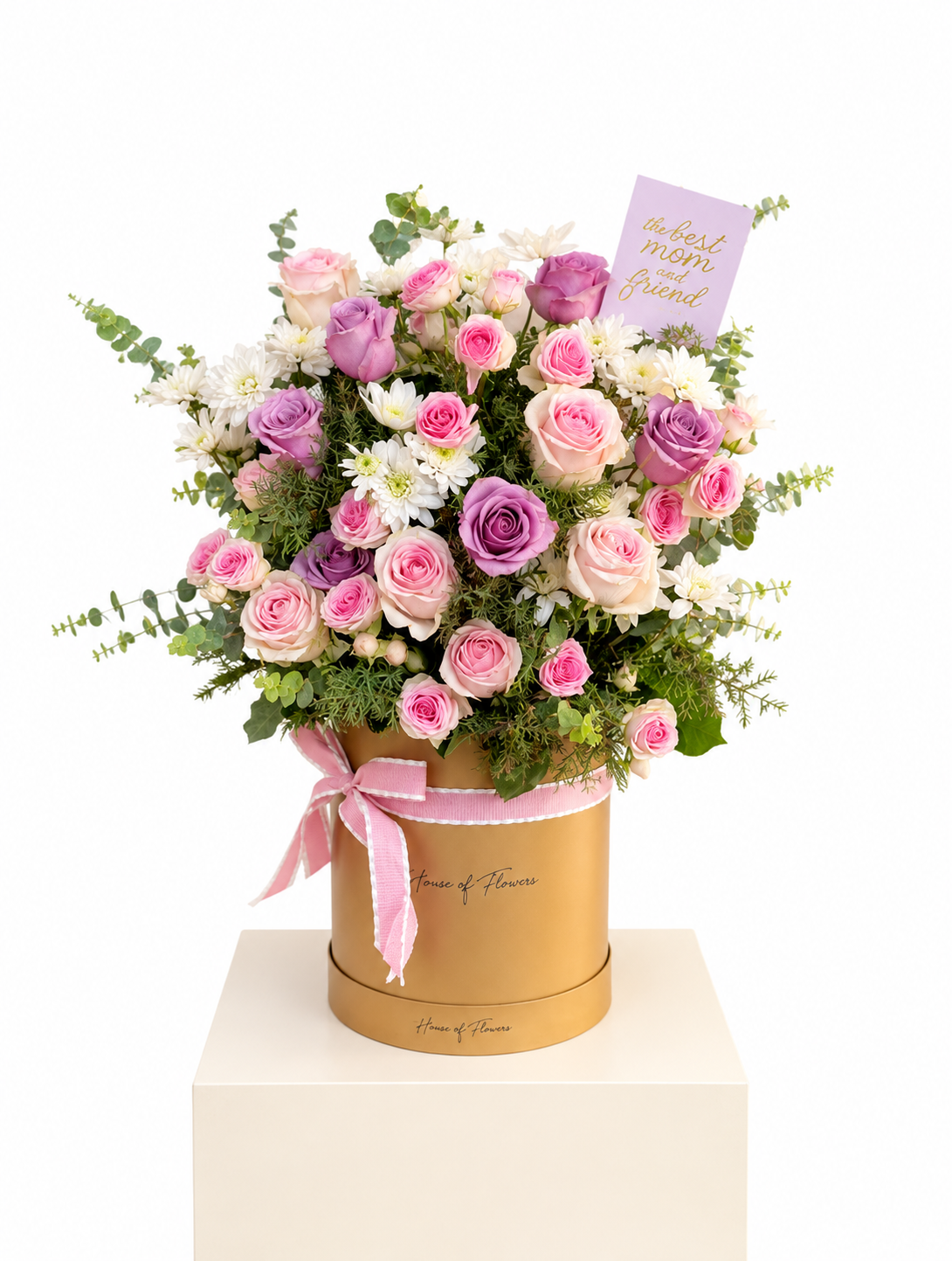 A floral arrangement of pink and purple roses, white daisies, and green foliage presented in a round gold gift box with a pink ribbon, on a white pedestal against a white background.