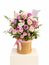 A floral arrangement of pink and purple roses, white daisies, and green foliage presented in a round gold gift box with a pink ribbon, on a white pedestal against a white background.