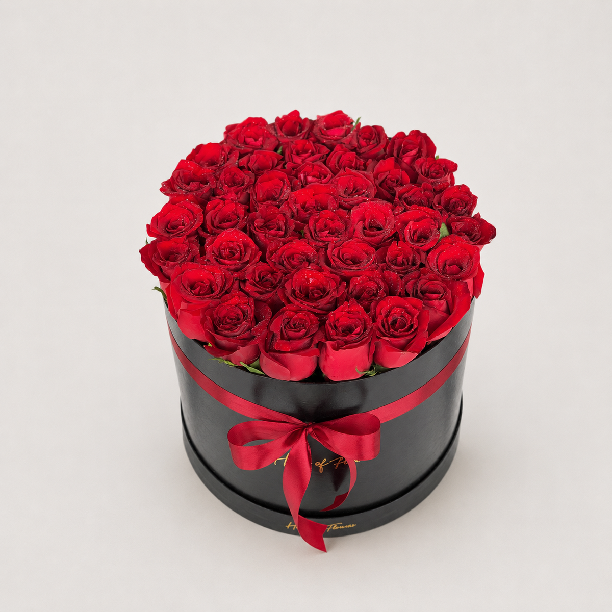 A round black gift box filled with 60 vibrant red roses, tied with a red satin ribbon and bow, set against a plain light gray background.