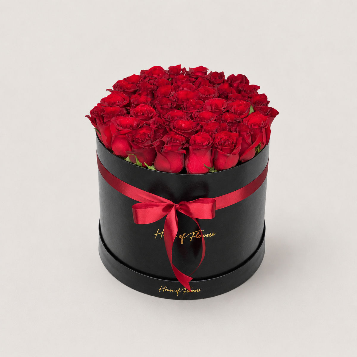 A round black gift box filled with red roses, tied with a red ribbon and bow, on a light gray background. The box has gold script branding that reads &