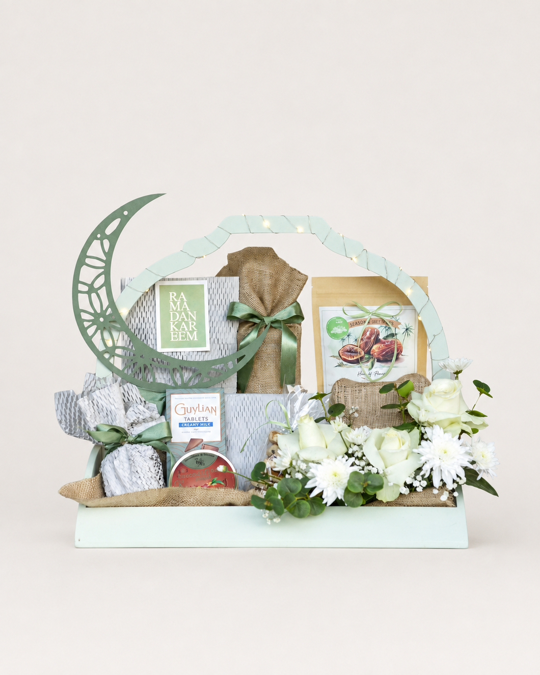 House of Flowers Soft Meadow Ramadan gift hamper in a mint green tray with crescent moon ornament, Guylian Creamy Milk chocolates, jute-wrapped Khatai, dry fruit pouch, Cavendish &amp; Harvey tin, Ramadan Kareem card, white roses, chrysanthemums, baby&