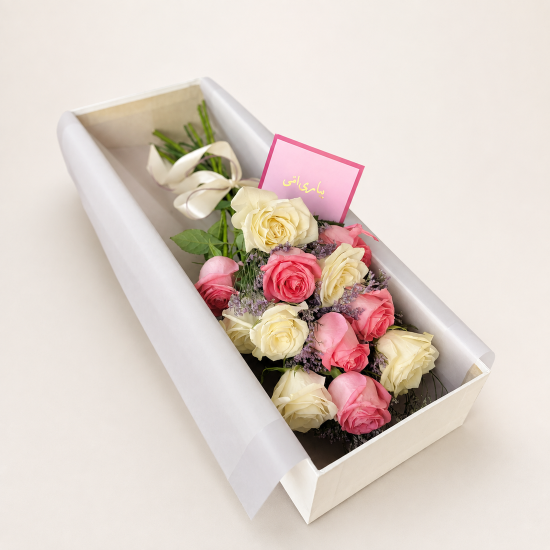 A gift box of roses containing pink and cream-colored roses, green stems, and a pink card with gold text, presented in a white box on a light beige background.