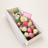 A gift box of roses containing pink and cream-colored roses, green stems, and a pink card with gold text, presented in a white box on a light beige background.
